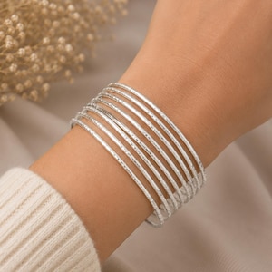 Puede incluir: Un conjunto de ocho brazaletes finos de metal plateado usados juntos. Cada brazalete presenta una superficie finamente texturizada que atrapa la luz, creando un sutil efecto brillante en la pila de pulseras circulares en la muñeca.