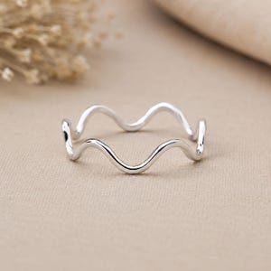 Puede incluir: Un anillo de plata pulida que presenta un diseño ondulado y serpenteante continuo. La banda tiene un acabado metálico liso y reflectante, con un grosor constante en toda su circunferencia, creando una forma ondulada simple y uniforme.