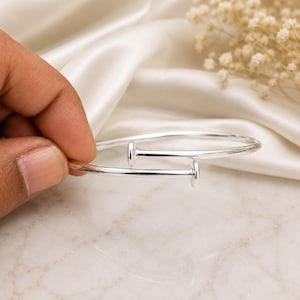 May include: A silver-toned metal bangle bracelet shaped like a nail, featuring a smooth, polished finish. The design consists of a single continuous band that curves around to form a circular shape, with the two ends resembling flat nail heads.