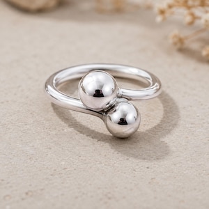 May include: A polished silver bypass ring featuring two smooth, spherical ball accents of different sizes at the open ends. The metal has a reflective, high-shine finish, and the band curves around to create a continuous, open-ended circular shape.