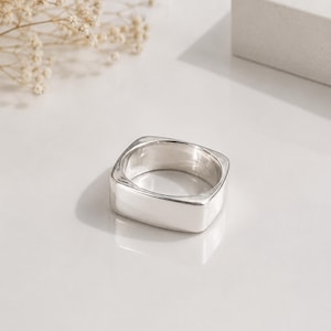 Op de afbeelding: Een gepolijste zilveren ring met een opvallende vierkante band. Het metalen oppervlak is glad en reflecterend, wat een strak, geometrisch ontwerp laat zien. De ring heeft een breed, plat profiel met afgeronde hoeken voor een solide uitstraling.