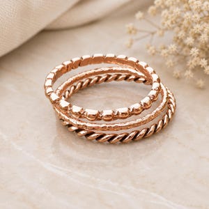 Op de afbeelding: Een set van drie gestapelde ringen in een roségouden tint. De eerste ring heeft een gladde kralentextuur, de tweede ring heeft een gehamerde afwerking en de derde ring vertoont een gedraaid touwontwerp. Elke ring is cirkelvormig en metaalachtig.