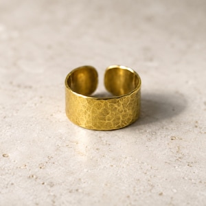 Peut inclure: Une bague ouverte et large de couleur dorée avec une surface martelée et texturée. L'anneau présente une largeur constante sur toute sa circonférence, avec une ouverture visible à l'arrière, révélant un éclat métallique et un aspect irrégulier.