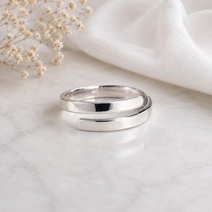 Può includere: Un anello bypass in tonalità argento lucido con un design a fascia liscia e aperta. L'anello presenta una finitura metallica riflettente e una struttura continua che si sovrappone, creando una silhouette semplice e pulita per il dito.