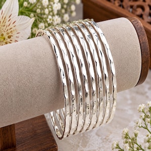 Pode incluir: Um conjunto de sete pulseiras rígidas de cor prateada com uma superfície martelada e texturizada. Cada pulseira tem um acabamento metálico reflexivo e uma forma circular consistente, apresentadas em pilha num suporte de joias.