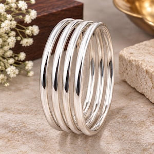 Peut inclure: Un ensemble de cinq joncs en métal argenté poli présentant un profil lisse et arrondi. Les bracelets sont disposés en pile, mettant en valeur une surface métallique réfléchissante avec des lignes épurées et une forme circulaire constante.