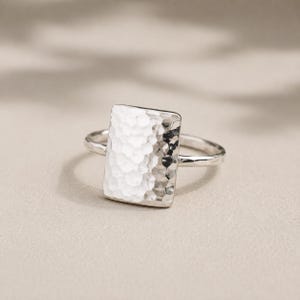 May include: A silver ring featuring a rectangular face with a hammered, textured surface. The band is thin and smooth, connecting to the sides of the rectangular centrepiece. The metal has a bright, reflective finish that catches the light.