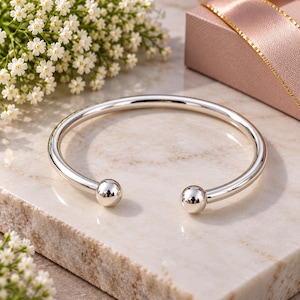 Può includere: Un bracciale rigido in argento lucido caratterizzato da una fascia circolare liscia aperta sul davanti. Le due estremità del bracciale sono rifinite con piccole sfere d'argento, creando un aspetto metallico pulito e riflettente.