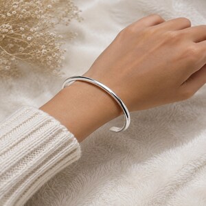 May include: A polished silver cuff bracelet with a smooth, rounded surface and an open-ended design. The bracelet features a simple, continuous band that encircles the wrist, showcasing a reflective metallic finish and a clean, minimalist aesthetic.