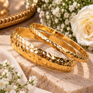 Puede incluir: Dos brazaletes dorados con una textura martillada y con hoyuelos en toda la superficie. Las pulseras son circulares, con una banda notablemente más ancha que la otra, ambas con un acabado metálico pulido y reflectante en su totalidad.