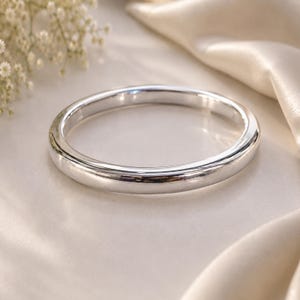 Pode incluir: Uma pulseira tipo bracelete de tom prateado polido com uma superfície lisa e arredondada e um design circular contínuo. O metal reflete a luz, criando reflexos brilhantes ao longo das bordas curvas da estrutura sólida da banda.