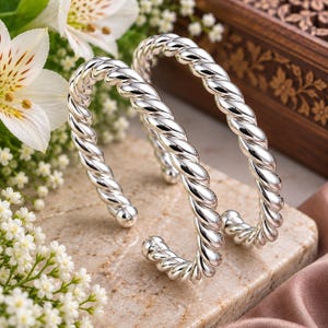Può includere: Due bracciali a fascia color argento con un design a corda intrecciata. Ogni bracciale ha una finitura lucida e riflettente con estremità arrotondate. I bracciali sono aperti per consentire la regolazione e presentano un motivo a spirale uniforme.