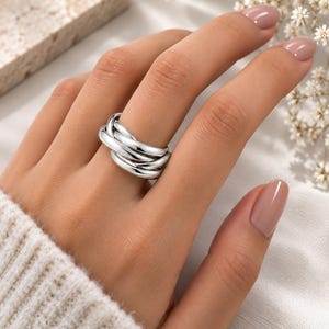 Peut inclure: Une bague en métal argenté poli présentant un design multi-anneaux composé de plusieurs boucles arrondies, lisses et interconnectées qui se chevauchent pour créer un aspect large et superposé. Le métal a une finition brillante et réfléchissante.