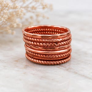 Peut inclure: Une pile de cinq bagues en cuivre présentant des textures alternées, incluant des anneaux martelés lisses et des anneaux torsadés en forme de corde. Les bagues sont empilées verticalement, révélant la finition métallique brun-rouge du cuivre.