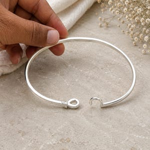 May include: A silver-toned open cuff bracelet featuring a smooth, polished finish. One end of the bracelet terminates in a small, circular loop wrapped with fine wire detailing, while the opposite end features a simple, curved hook design.