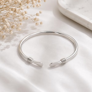 May include: A polished silver cuff bracelet featuring a smooth, open-ended design. Each end of the bracelet is accented with three delicate, raised horizontal wire wraps, creating a clean and reflective metallic finish throughout the piece.