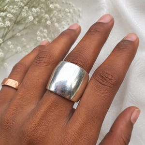 Puede incluir: Un anillo de banda ancha en tono plateado pulido con una superficie lisa y curva. Al lado, se observa un anillo de banda más fina en tono cobre con textura. Ambos anillos presentan un acabado metálico y un diseño sencillo sin adornos.