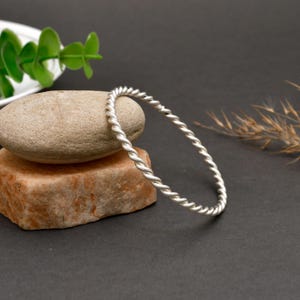 Può includere: Un bracciale rigido color argento caratterizzato da una texture a corda ritorta continua. Il cerchio metallico presenta una finitura opaca e uno spessore uniforme lungo tutto il design, creando un'estetica semplice e pulita per il polso.