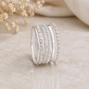 May include: A set of five silver rings stacked together. The collection includes a plain band, a twisted rope band, a band with floral engraving, and two bands featuring small, clear, circular stones set along the entire circumference of the metal.
