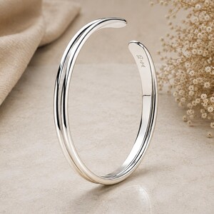 May include: A polished silver cuff bracelet with a smooth, rounded profile and an open design. The inner surface of the bracelet features a clear 925 hallmark stamp, indicating sterling silver composition, along with a small decorative maker's mark.