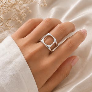 Könnte beinhalten: Ein silberfarbener Ring mit einer quadratischen Vorderseite und einer kreisförmigen Aussparung in der Mitte. Die Ringschiene ist glatt und poliert und zeigt ein geometrisches Design, das flach am Finger anliegt. Das Metall hat eine reflektierende Oberfläche.