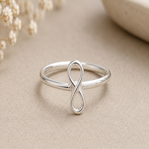 May include: A polished silver ring featuring a vertical infinity symbol design centred on a smooth, thin band. The metal has a reflective, bright finish, and the infinity loop is formed by a continuous, slender wire that creates a balanced, symmetrical shape.