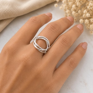 May include: A polished silver ring featuring a multi-band interlocking design. The ring consists of three thin, smooth, circular bands that are looped together to create a continuous, layered appearance on the finger, showcasing a bright metallic finish.