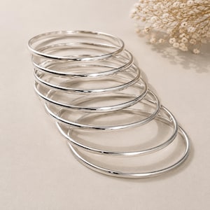 May include: A set of seven polished silver-colored bangle bracelets arranged in a cascading stack. Each bangle features a smooth, rounded, and reflective surface with a consistent circular shape, creating a uniform appearance across the entire collection.