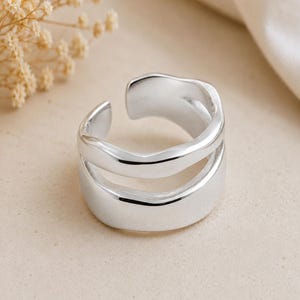 Puede incluir: Un anillo de tono plateado pulido con un diseño abierto de doble banda y bordes suaves y ondulados. El anillo presenta una estructura ancha de vástago dividido que crea un espacio entre las dos bandas, mostrando un acabado metálico reflectante.