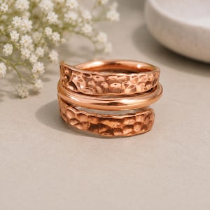 May include: A rose gold-toned wrap ring featuring a multi-layered design. The ring consists of two outer bands with a hammered, textured finish and a single smooth, polished central band, creating a stacked appearance in a continuous spiral.