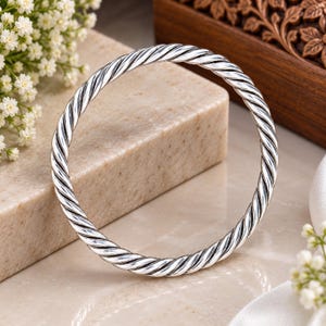 Puede incluir: Un brazalete circular de plata con un diseño continuo de cuerda trenzada. La superficie metálica tiene un acabado pulido con áreas hundidas oscurecidas entre las torsiones para resaltar la intrincada textura en espiral de la banda metálica.