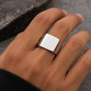 Könnte beinhalten: Ein polierter silberfarbener Siegelring mit einer glatten, flachen, quadratischen Oberfläche. Der Ring hat eine massive, breite Ringschiene, die sich nach hinten leicht verjüngt und eine saubere, reflektierende Metalloberfläche ohne Gravuren zeigt.