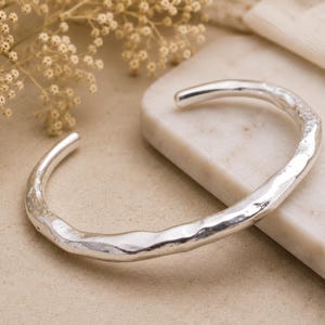 May include: A silver cuff bracelet with an open design and a textured, uneven surface. The metal has a polished, reflective finish that catches the light, highlighting the organic, undulating shape of the band as it curves into a circular form.