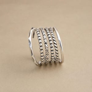 Könnte beinhalten: Ein silberner Mehrfachring bestehend aus fünf verschiedenen, übereinanderliegenden Segmenten. Das Design umfasst einen glatten Ring, einen Perlenring, einen Ring mit X-Muster, einen weiteren Perlenring und einen gedrehten Kordelring, die alle verbunden sind.