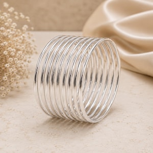 Può includere: Un bracciale a fascia composto da dieci sottili anelli circolari color argento lucido, collegati tra loro per formare un unico polsino largo. La superficie metallica riflette la luce, creando riflessi luminosi lungo la struttura liscia e arrotondata del filo.