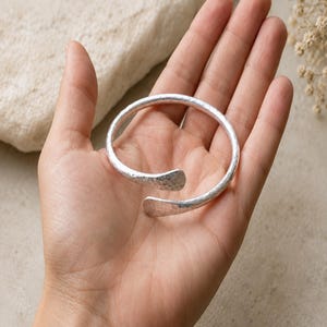 May include: A silver-colored open cuff bracelet with a textured, hammered finish. The bracelet features a circular band that does not fully close, ending in two slightly flattened, rounded terminals. The metal surface reflects light across its uneven, dimpled pattern.