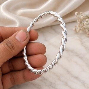 Può includere: Un bracciale rigido in argento lucido caratterizzato da un design a corda intrecciata continua. Il cerchio metallico presenta una superficie liscia e riflettente con un motivo a spirale uniforme che avvolge l'intera circonferenza del gioiello.