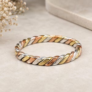 Puede incluir: Un brazalete rígido tricolor con un diseño de cuerda trenzada. La pulsera está construida con tres hilos metálicos distintos en tonos oro amarillo, oro rosa y plata, entrelazados para crear una banda circular continua.