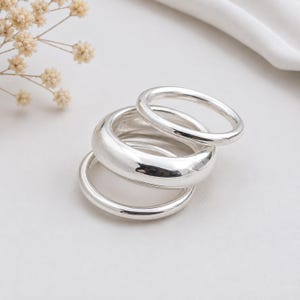 May include: A set of three polished silver rings stacked together. The rings feature smooth, rounded bands of varying thicknesses, with one wider band positioned between two thinner, uniform bands, all displaying a bright, reflective metallic finish.