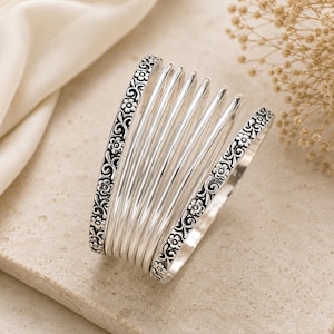 May include: A multi-strand silver cuff bracelet featuring two wide outer bands with an embossed floral and scroll pattern and several smooth, rounded inner bands that create a layered, stacked appearance with a polished metallic finish.