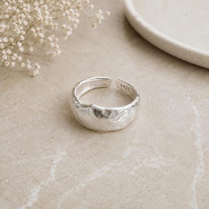 Puede incluir: Un anillo de plata con un diseño de banda abierta y ajustable que presenta una superficie texturizada e irregular. El interior de la banda está claramente marcado con el texto S925, indicando la composición metálica de la pieza. Acabado metálico.