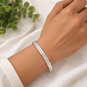 Könnte beinhalten: Ein polierter Silberarmreif mit einem eleganten, mehrsträngigen Design, bei dem zwei parallele Metallbänder durch kleine, zylindrische Silberakzente an den Seiten verbunden sind, was ein klares und einheitliches Erscheinungsbild am Handgelenk erzeugt.