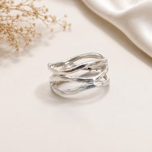 May include: A silver ring featuring a multi-layered, open-work design with three distinct, wavy, and textured bands that overlap and connect to form a single cohesive piece of jewellery with a polished, reflective metallic finish.