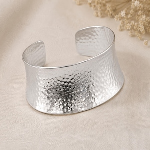 Pode incluir: Uma pulseira larga de prata com uma textura martelada distinta em toda a sua superfície externa. O metal tem um acabamento polido e reflexivo com um design curvo e aberto que mantém uma largura constante em toda a peça.