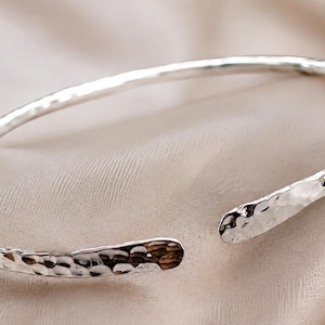 Könnte beinhalten: Ein silberfarbenes offenes Armband mit einer strukturierten, gehämmerten Oberfläche über das gesamte Band. Das Armband hat eine kreisförmige Form mit zwei abgerundeten, leicht abgeflachten Enden, die sich nicht berühren und eine Lücke bilden.