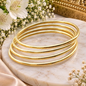 Puede incluir: Un conjunto de cuatro brazaletes finos de metal dorado pulido con una superficie lisa y redondeada. Las pulseras están apiladas juntas, creando una forma circular con un acabado metálico reflectante que capta la luz en sus bandas.