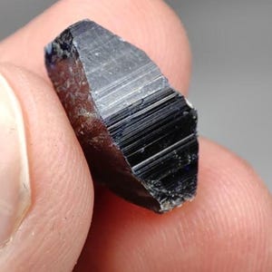 May include: A raw, dark blue to black crystal specimen with a distinct elongated hexagonal shape and prominent vertical striations along its faceted sides. The mineral displays a glossy, reflective surface texture with sharp, angular edges and a deep, opaque coloration throughout its structure.