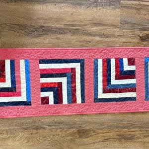 May include: A pink quilted table runner with four square patches of fabric. Each patch is made of red, white, and blue fabric strips sewn together in a geometric pattern.