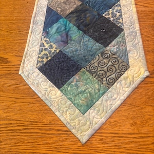 May include: A quilted table runner featuring a central panel of diamond-shaped fabric patches in various shades of blue, teal, and grey. The border is a light cream fabric with a continuous machine-stitched swirl pattern throughout the edges.
