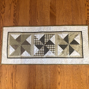May include: A rectangular quilted table runner featuring three pinwheel designs. The pinwheels are made of brown, white, and olive green fabrics, including gingham and solid patterns. The runner has a white border with a decorative edge.